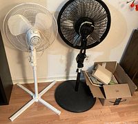 Two standing fans shown side by side, one white and one black with remote, next to a box