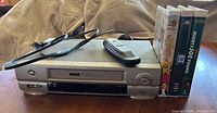 Overall view of the RCA VCR player with power cord, black remote control, and three VHS movie cases laid out on a fabric surface.