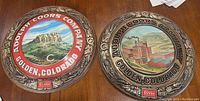 Two vintage round Coors plaques on a table, side by side, showing distinct brewery artwork under glass with decorative rims