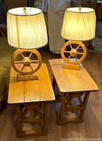 Photo of two wooden wagon wheel side tables side by side, each supporting a wagon wheel lamp with a lit shade, showing overall condition and design.