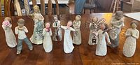 Wide front view of all 11 Willow Tree figurines arranged on a wooden table showing various figures including angels and children with several holding objects like hearts, flowers, or a bird.