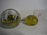 Two glass paperweights shown side by side. The left one is spherical with a hollow rounded center and bubble design. The right one is a solid clear glass sphere with an internal yellow floral pattern.
