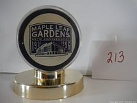 Front view of the commemorative Maple Leaf Gardens 65th anniversary hockey puck in clear plastic display case mounted on a gold-colored metal stand.