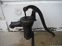 Front angle view showing the cast iron body, manual lever handle, and spout of the antique well pump. Visible wear and rust.