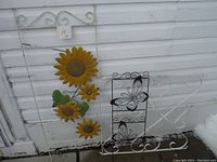 Three metal garden trellises leaning against a wall. One decorated with yellow sunflowers, one with black metal butterflies, all with white scrollwork frames.