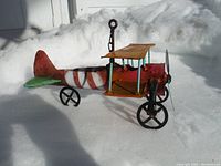 Side view of metal airplane sculpture on snow, showing red and white striped fuselage, green tail wings, orange upper wings with rust, two black wheels, blue propeller and support rods, and hanging loop with chain on top.
