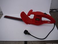 Side view of the electric hedge trimmer showing red plastic body, handle, safety guard, and attached power cord with plug.