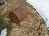 Side view of rusted metal lawn roller lying on grass showing cylindrical drum and red metal handle