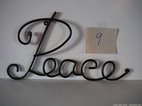 Front view of the black metal 'Peace' word wall art lodged on a white background with a sticky note labeled '9' nearby.