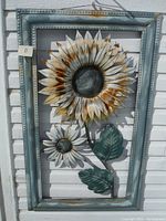 Full view of the large metal sunflower garden art piece in a blue-gray riveted frame hanging on white siding, showing the rust spots and overall structure.