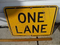Front view of the yellow metal sign with 'ONE LANE' in black text, showing wear and age marks.