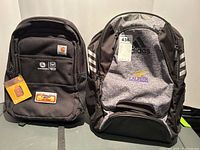 Both backpacks shown side by side. The Carhartt backpack is black with logos and patches, appearing brand new with tags. Adidas backpack is grey and black with Laurier logo and Adidas branding, gently used.