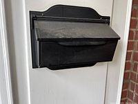 Black mailbox mounted on white wall, closed top lid, dust visible on surface.