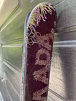 Single ski angled against garage door showing burgundy top sheet with stylized yellow/pink floral motif and white Cantika graphics