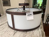Photo of ceiling light fixture showing side view with white frosted shade and dark brown frame accents, including round mounting piece on top.