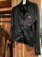 Front view of black The North Face Summit Series men's jacket hanging on a wooden hanger, showing full length with zip up and chest pocket with logo.
