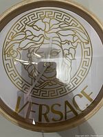 Photo of a round wall clock featuring the Versace Medusa head logo and the name 'VERSACE' in gold on a white background with a gold frame.