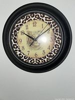 Image of round wall clock with a tiger head in the center and a leopard print border pattern, set within a black frame.