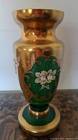 Full vase front view showing green glass, gold plating, hand painted white floral design with gold scrollwork.