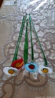 Four art glass flowers and one green glass leaf with long stems laid out on a beige lace textured cloth.