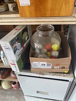 Jar filled with several dozen golf balls in white, neon yellow and orange colors beside a boxed electric putting green.