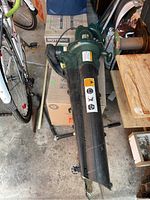 Full view of Yardworks electric leaf blower showing green motor housing and black blower tube, placed on a box with other garage items around.