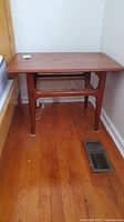 Full view of the teak table against a wall near a bed and floor vent, showing the overall shape and cane shelf below.