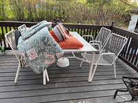 Full view of the patio set on wooden deck. Includes 4 white metal wire frame chairs, white slatted metal table, orange and patterned cushions, and the umbrella resting on chairs.