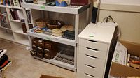 White plastic shelving unit with three shelves below a top surface, displaying organized items underneath and next to it in a room.