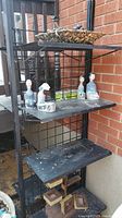 Metal shelving unit with four flat shelves and wire grid sides placed outdoors next to brick wall and wooden railing. Some decorative items visible on shelves.