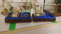 Two silver bicycle driver figures displayed in glass cases on decorative bases with blue fabric lining.