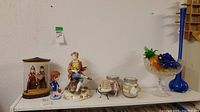 Photo showing the complete set of items on a shelf including the King Rama and Queen Shinta figurines in case, bobblehead doll, boy figurine with missing arm, glass fruit bowl, tall blue vase, and candles.