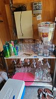Wide view of the beverage dispenser and multiple acrylic drinking glasses, tumblers, stemware, and stirrers on a metal utility cart in a shed.