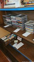 Wooden shelf with multiple stacks of music CDs in jewel cases and a box underneath with cassettes and paper notes.