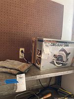 Photo showing the circular saw in original box on shelf alongside a pair of beige work gloves with green trim, against a pegboard background.