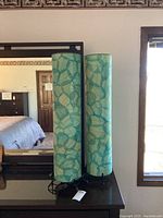 Pair of tall cylindrical lamps with teal and blue dyed cocoa leaf shades on tops of black metal circular bases placed on a dark dresser in front of a mirror.
