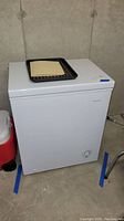 Front view of white chest freezer with black tray and envelope on top, placed on concrete floor.