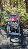 Front view of the Hose Mobile hose reel with coiled white hose and red plastic bin behind it on asphalt ground by a chain link fence.