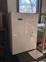 Front and side angled view of white Haier mini fridge on a dark table near a window in a garage setting, revealing overall condition and size.