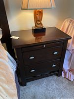 Straight front view of dark wood nightstand showing two drawers with silver oval pulls, pull-out shelf, and lamp on top.