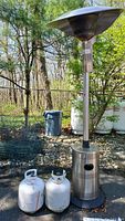 Photo showing portable propane patio heater with two standard propane tanks set outdoors on concrete with wooded background.