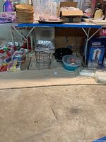 Photo of a blue folding table with stacked wicker placemats, clear plastic party cups, metal wire racks, black trays, plastic bowls in blue, and boxed DeLonghi coffee maker under the table.