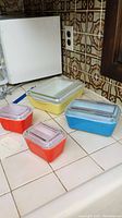 Five vintage Pyrex refrigerator storage boxes in yellow, red, blue, and purple-tinted colors with clear lids on white tiled counter.