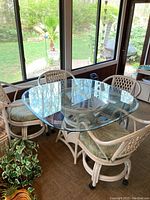 Square glass top rattan table with four matching rolling chairs with cushions surrounding it, situated inside a room with large windows overlooking a green yard.
