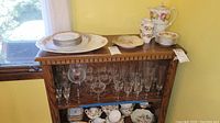 Wooden shelf displaying multiple glass stemware pieces inside, plates and platter on top shelf, tea set on bottom shelf.