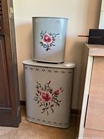 Two vintage metal enamelware pieces stacked: large hamper on bottom and smaller waste can on top. Both have pink and white floral hand-painted decorations on light gray background with decorative perforations.