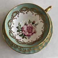 Top view of the tea cup showing the pink cabbage rose inside with gold filigree and turquoise lip, alongside the matching saucer with gilt filigree on turquoise.