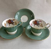 Two vintage Aynsley footed tea cups on matching sage-green saucers with gold rims and a saucer in the background.