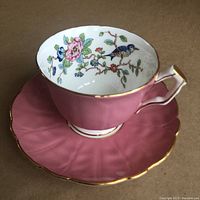 Photo shows pink footed teacup with detailed floral and blue bird interior design on a matching pink saucer with gold trim.
