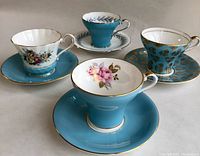Four Aynsley tea cups and their matching saucers arranged on a flat surface, showing varied blue exteriors and distinct patterned interiors.